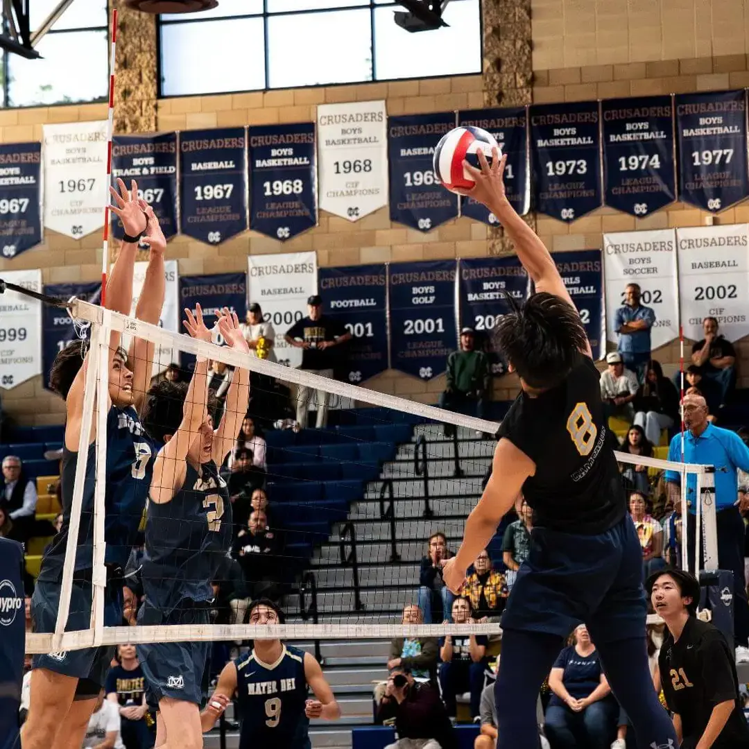 Cambridge upper school boys' volleyball team competes in a high-energy match, with one player spiking the ball as two opponents jump to block, showcasing teamwork and athletic excellence.