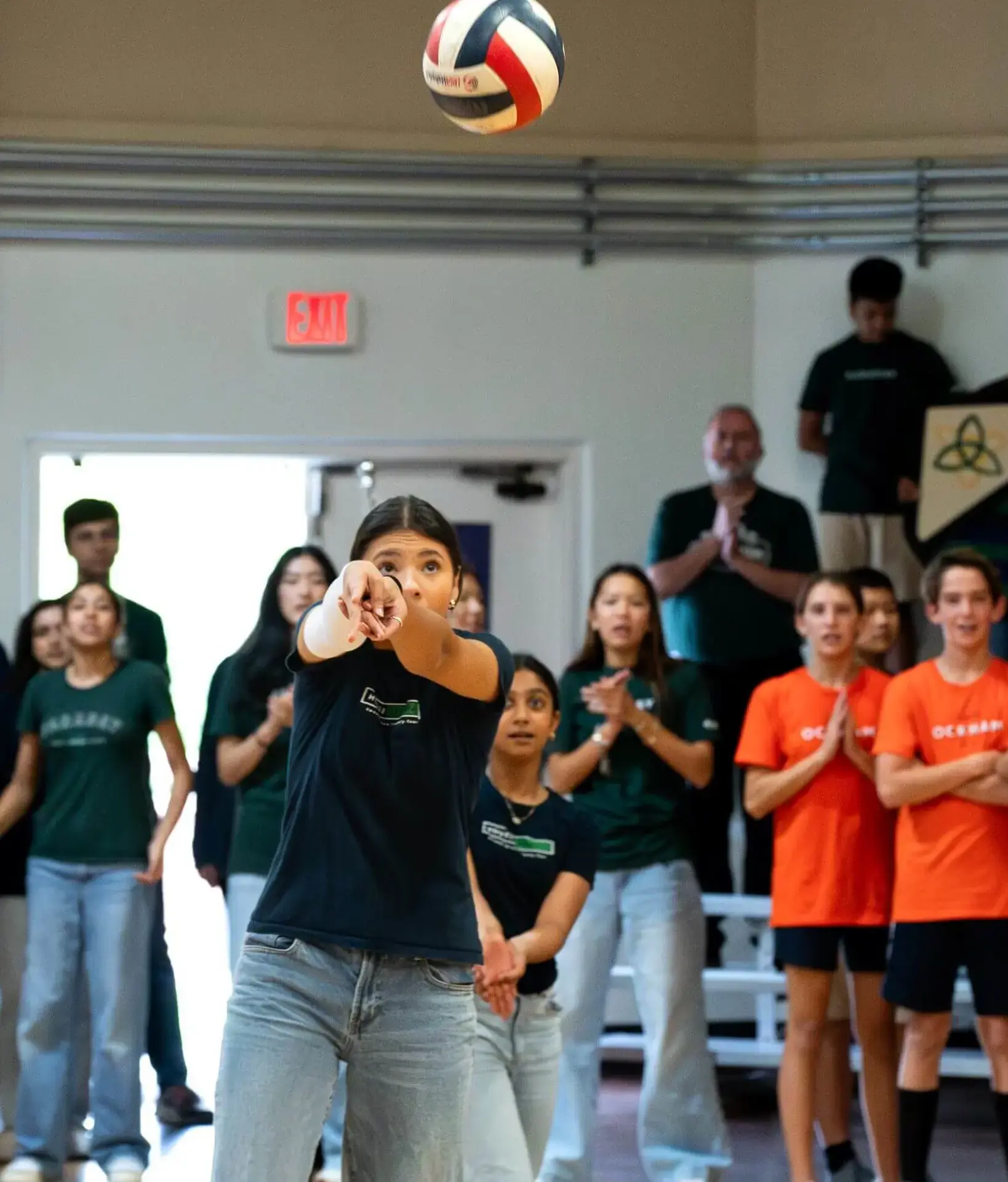 Upper school student aims to serve during a spirited volleyball match at a Cambridge house competition, while peers in green and orange house shirts cheer her on in the background.
