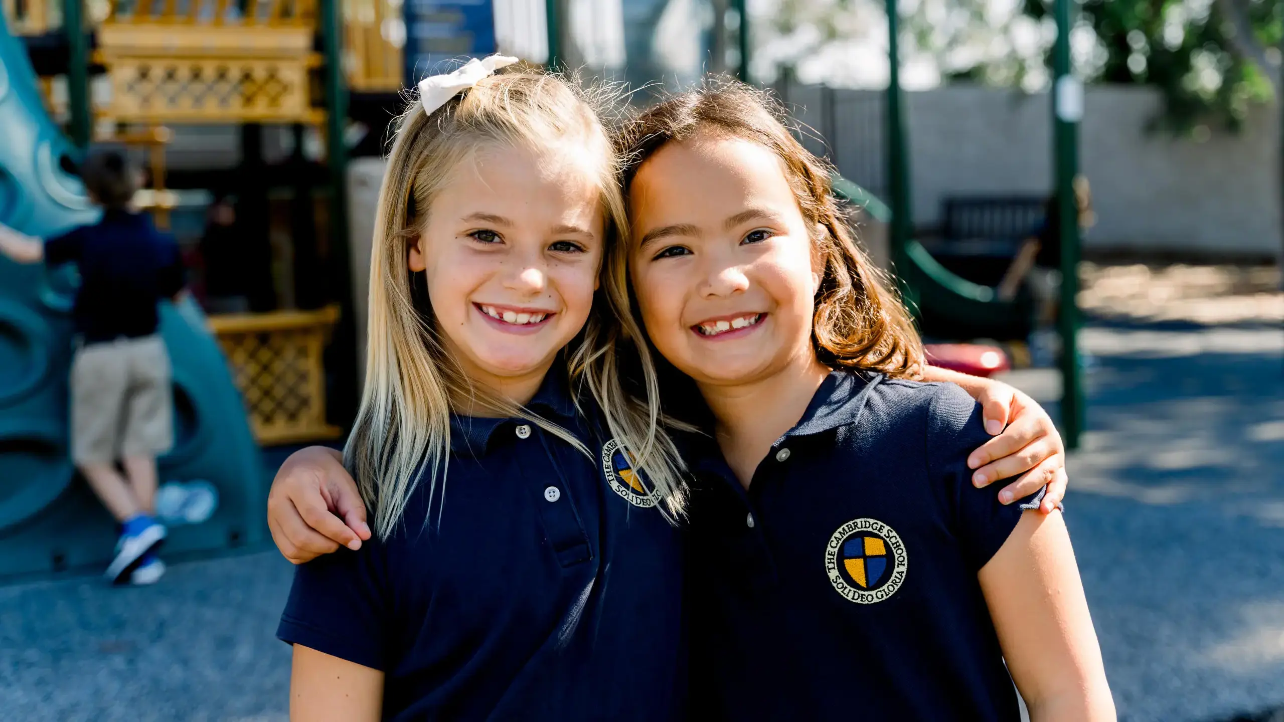 Two smiling elementary students from The Cambridge School embrace on a sunny playground, reflecting a nurturing and joyful learning environment where children flourish.
