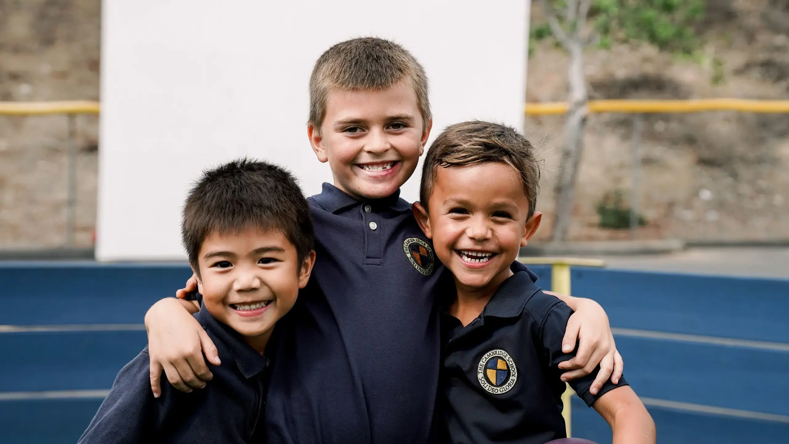 Three young boys in The Cambridge School uniforms smile and hug during recess at this private Christian school in San Diego, known for its joyful classical education and strong sense of community.