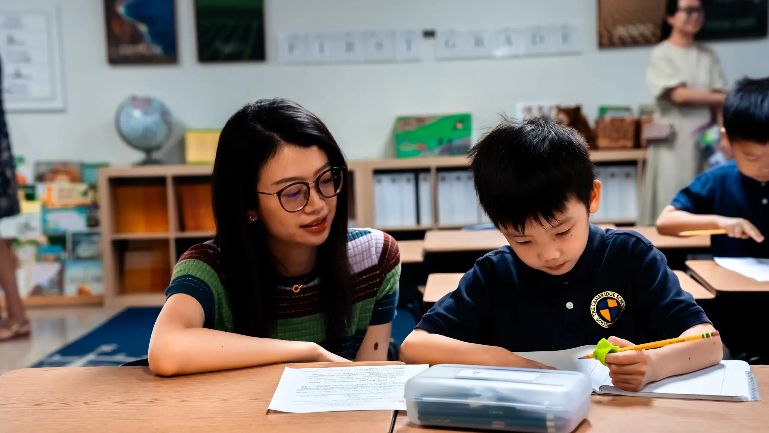 Inside a Cambridge School classroom, a parent attentively assists a student with their work.