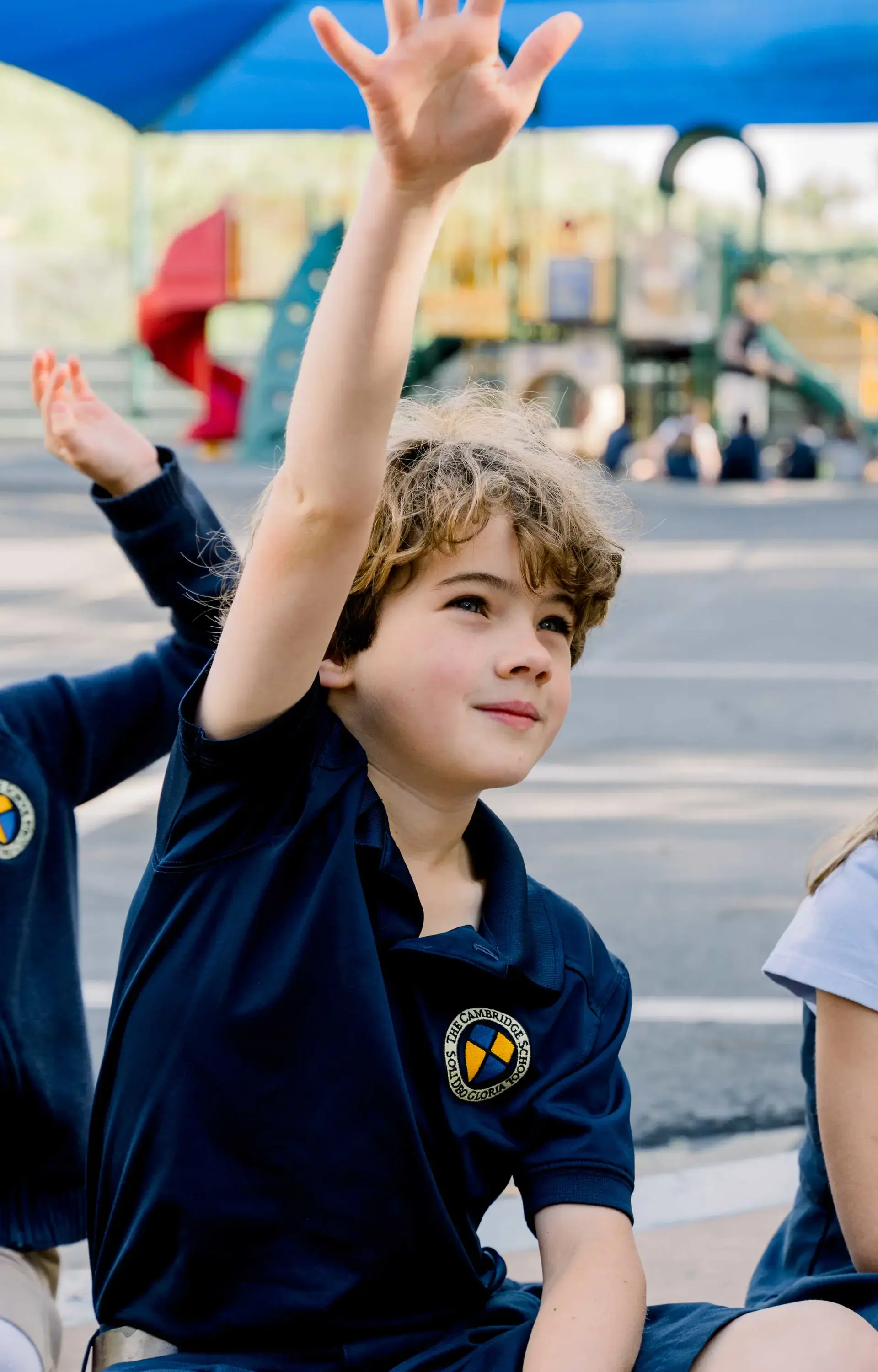 Young Cambridge School student in a navy polo eagerly raises his hand during an outdoor class session, reflecting curiosity and active participation in learning.