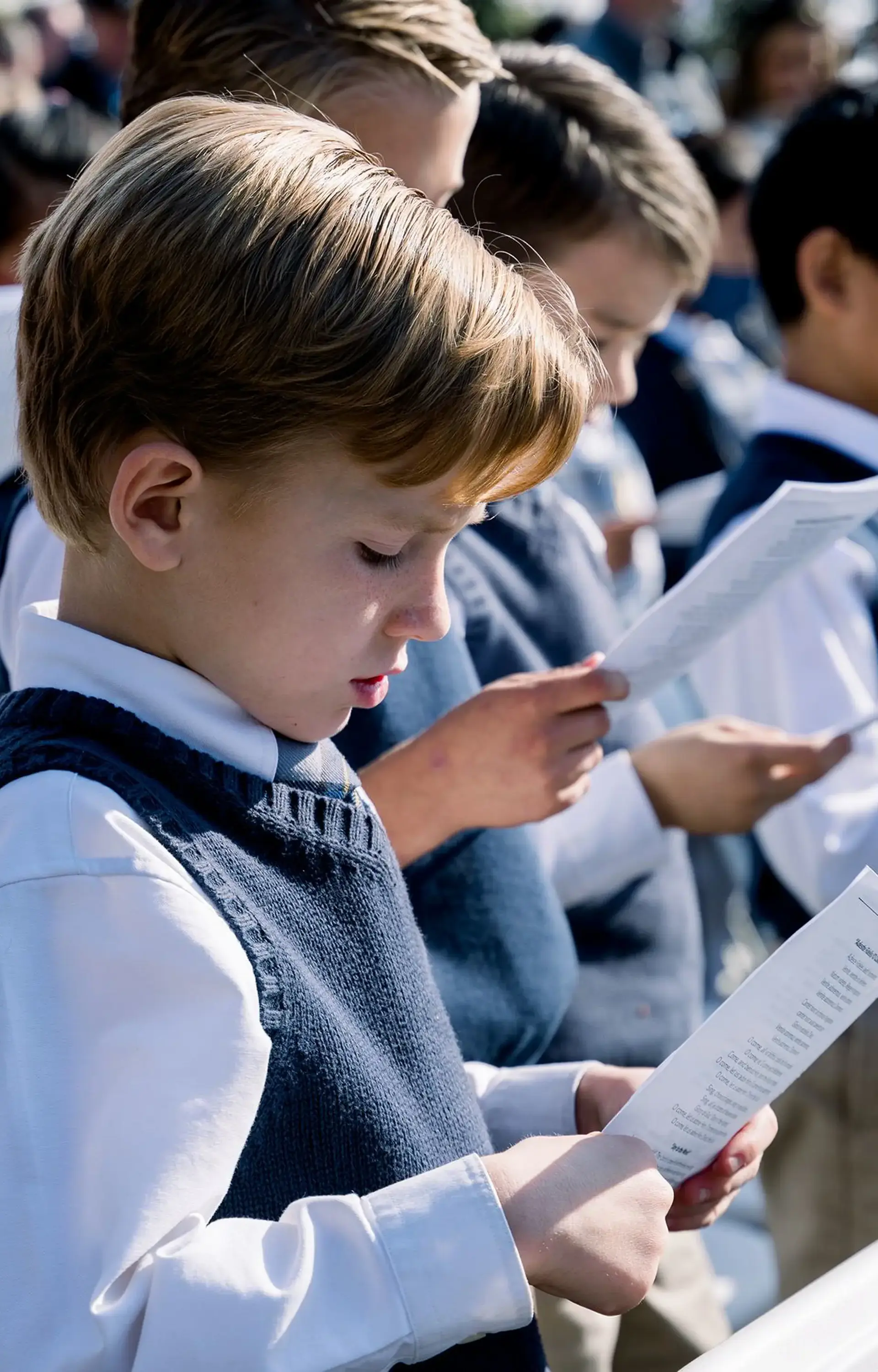 Young Cambridge School students in dress uniform sing from printed papers during an outdoor assembly or ceremony, with sunlight casting soft shadows across the group.